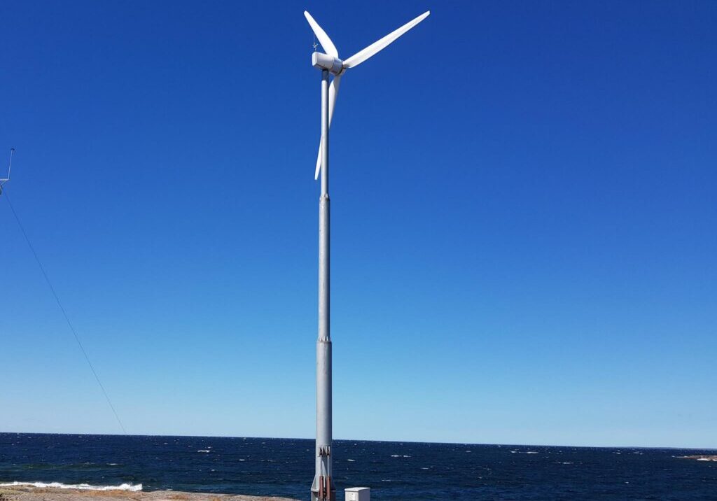 Moderni valkoinen pientuulivoimala asennettuna kallioiselle merenrannalle. Taustalla aukeaa sininen meri ja taivas.