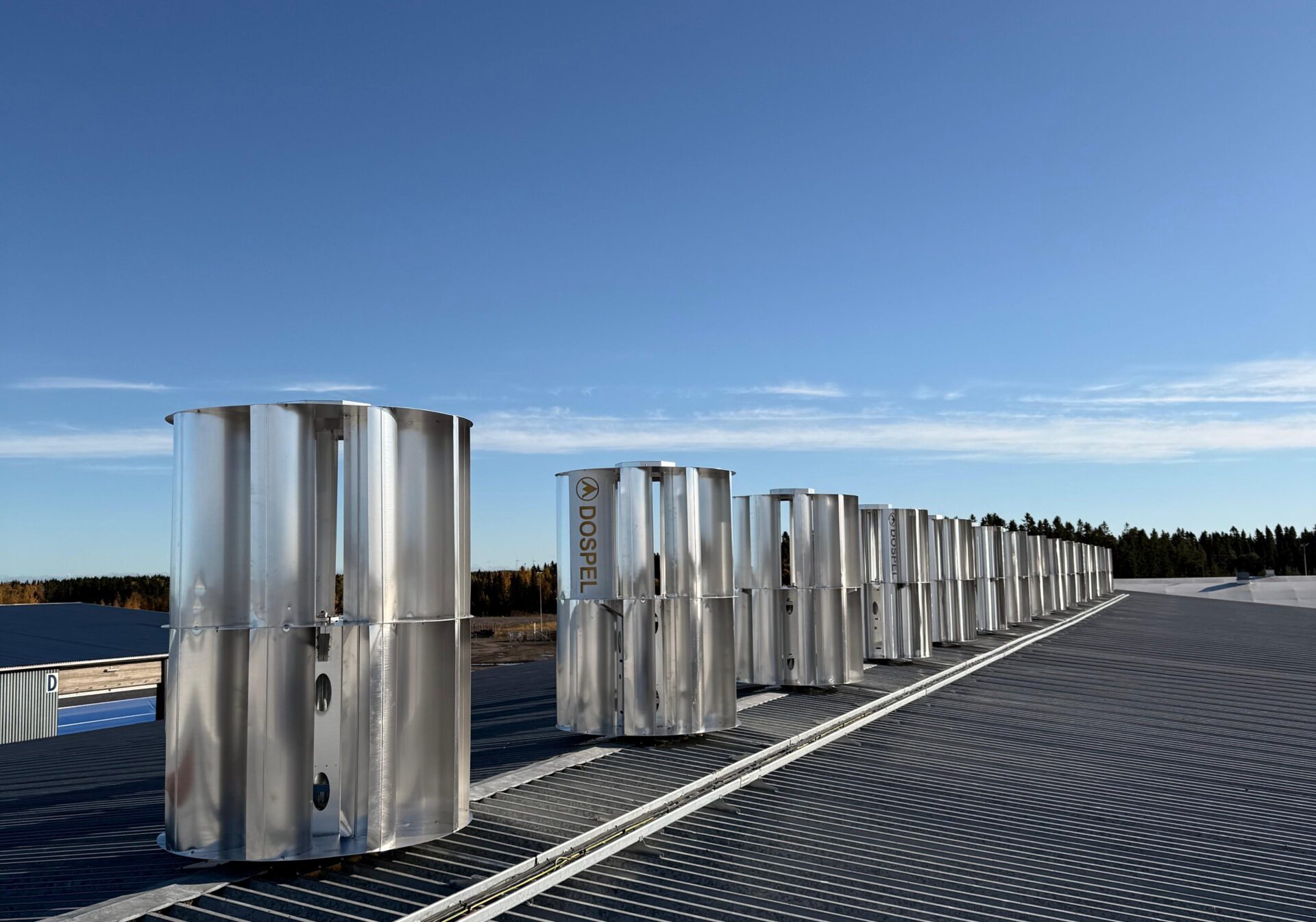 Pitkä rivistö hopeisia Dospel-pystyakselituulivoimaloita asennettuna suuren rakennuksen peltikatolle aurinkoisena päivänä.